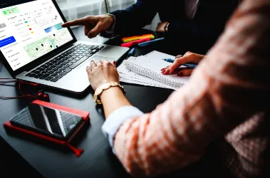 A man and woman sit at a table, focused on a laptop in front of them.